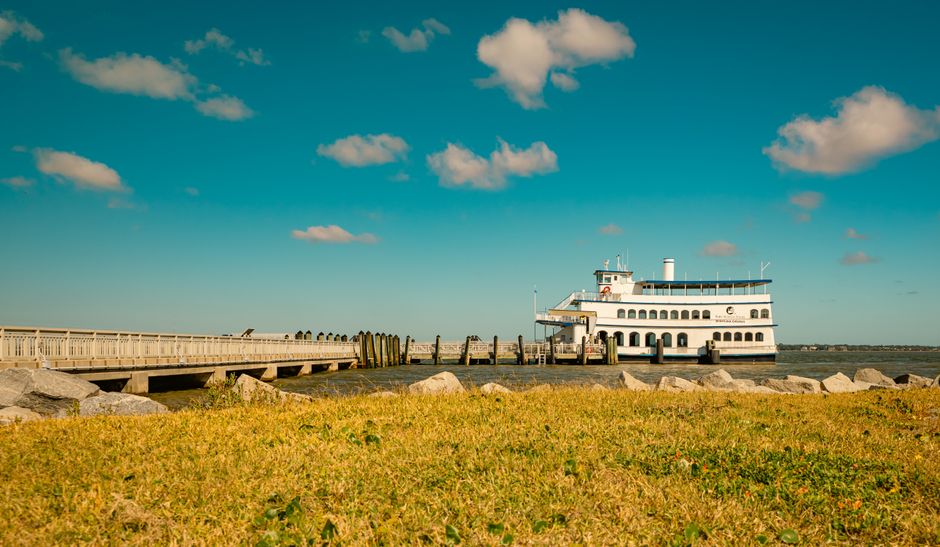 A large white and blue ferry vessel with the name 'HARD' docked at a harbor, approaching a wooden pier with rusted metal barriers. The ferry has multiple decks, with antennas, navigation equipment, an