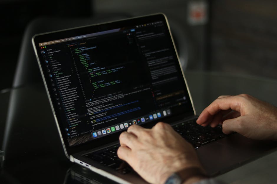 A close-up of a person's hand using a laptop with a silver body, positioned on a wooden surface. The screen displays lines of code with syntax highlighting, suggesting programming or web development a